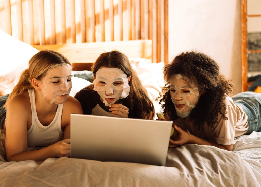 Drei Mädchen, mit Tuchmasken im Gesicht, liegen auf dem Bett und schauen auf einen Bildschirm